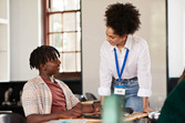 Student sitting at desk smiling up at staff standing, leaning on desk, appearing to provide support