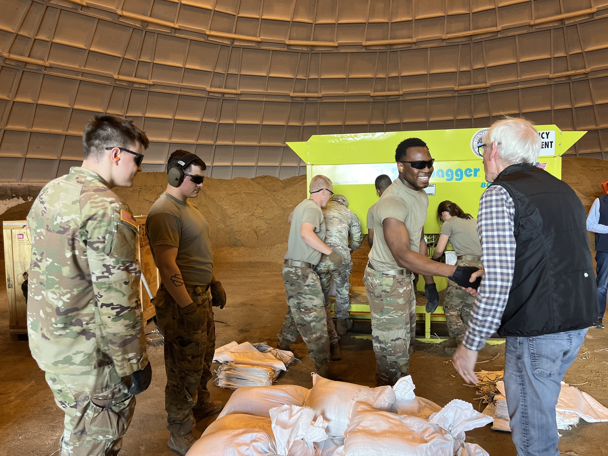 Governor Evers shakes hands with a member of the Wisconsin National Guard while other members of the Wisconsin National Guard pack sandbags. 