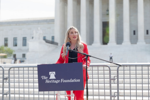 Megan Degenfelder stands grasping a podium wearing a red blazer and black blouse in front of the US Supreme Court Building.