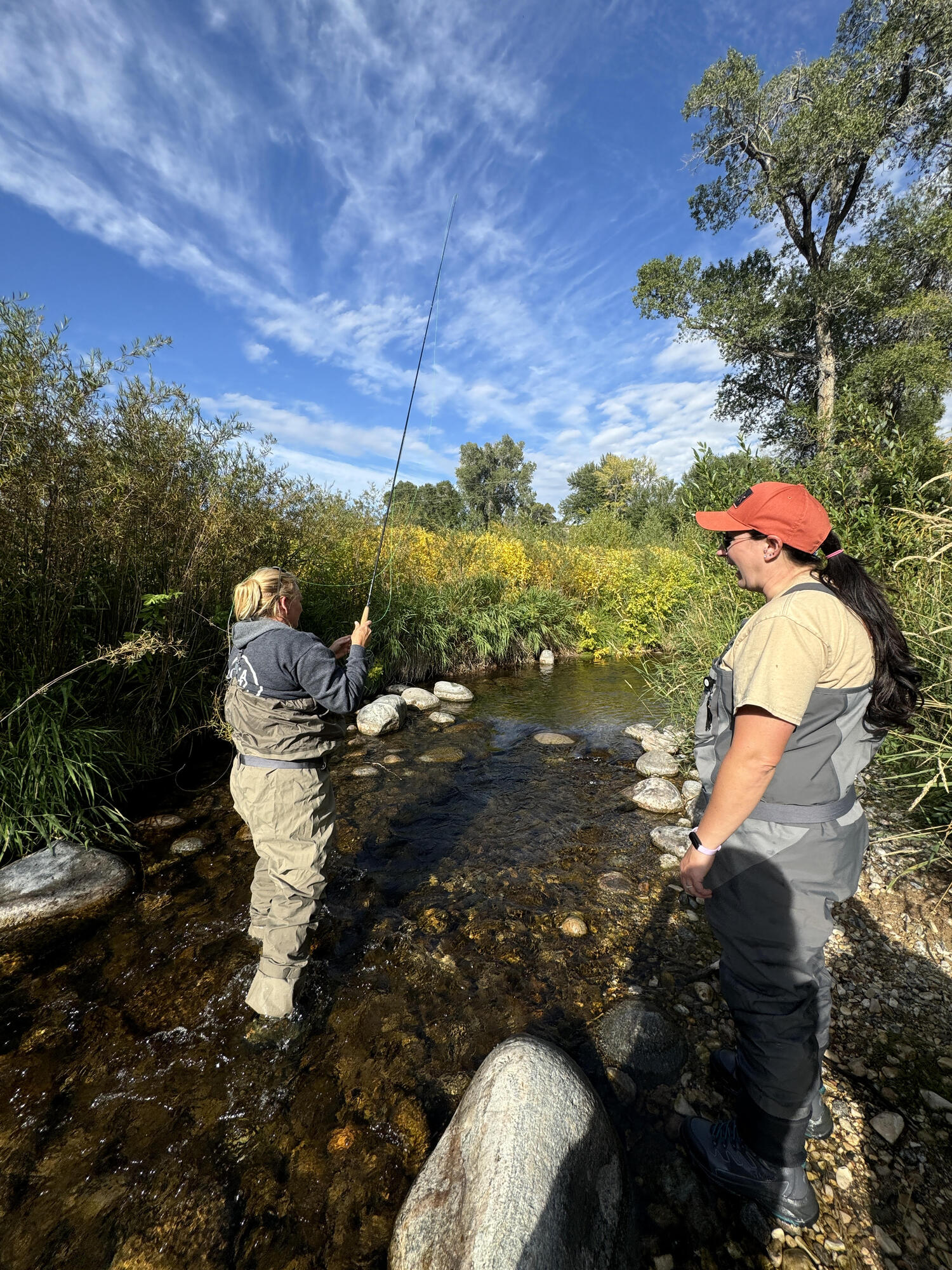 woman fly fishing with woman instructing, in a creek in waders